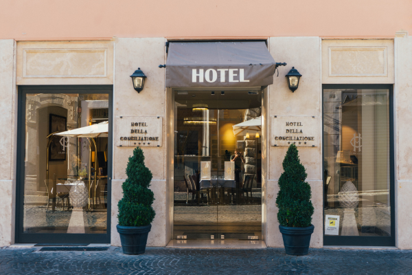 Entrada de hotel com fachada charmosa, duas plantas decorativas e toldo cinza, sugerindo uma hospedagem acolhedora e bem localizada