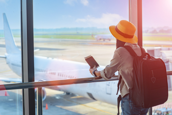 Mulher com chapéu e mochila observa avião pela janela de aeroporto, prestes a embarcar em uma viagem curta.
